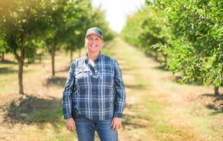 A person in a plaid shirt and cap stands smiling in the middle of a sunny orchard lined with ripe prune trees.