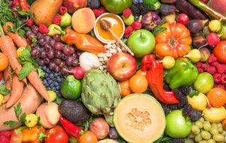 A variety of fresh fruits and vegetables, including carrots, apples, oranges, grapes, artichokes, peppers, berries, celery, and melons, displayed together in a colorful arrangement.