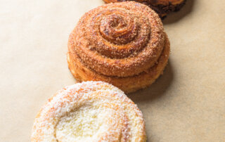 Three spiral pastries are arranged in a diagonal line on parchment paper, each with a different topping: chocolate, cinnamon sugar, and shredded coconut