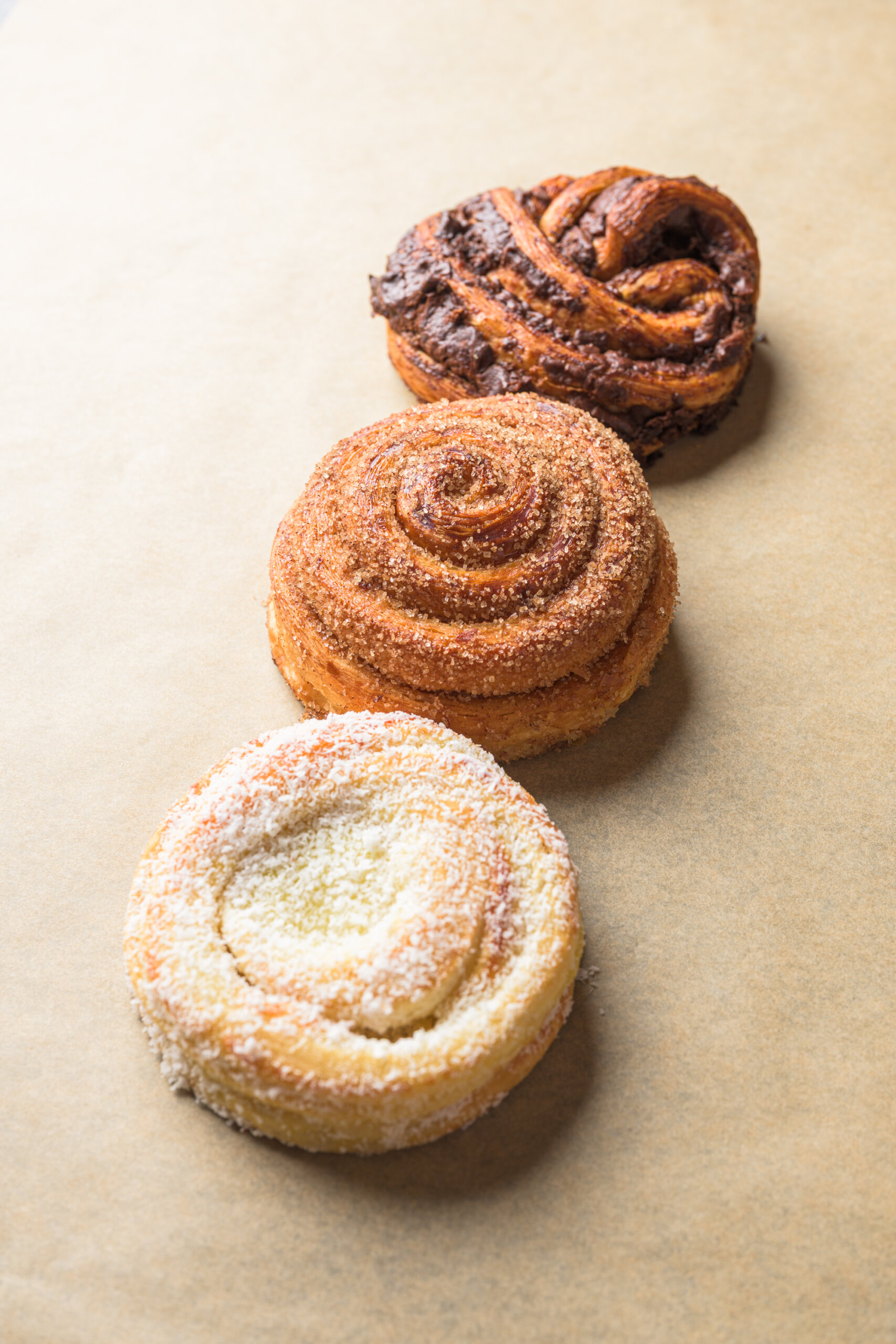 Three spiral pastries are arranged in a diagonal line on parchment paper, each with a different topping: chocolate, cinnamon sugar, and shredded coconut