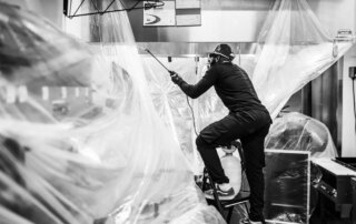 A person stands on a step ladder, spraying liquid in a kitchen area covered with plastic sheets for cleaning or maintenance.