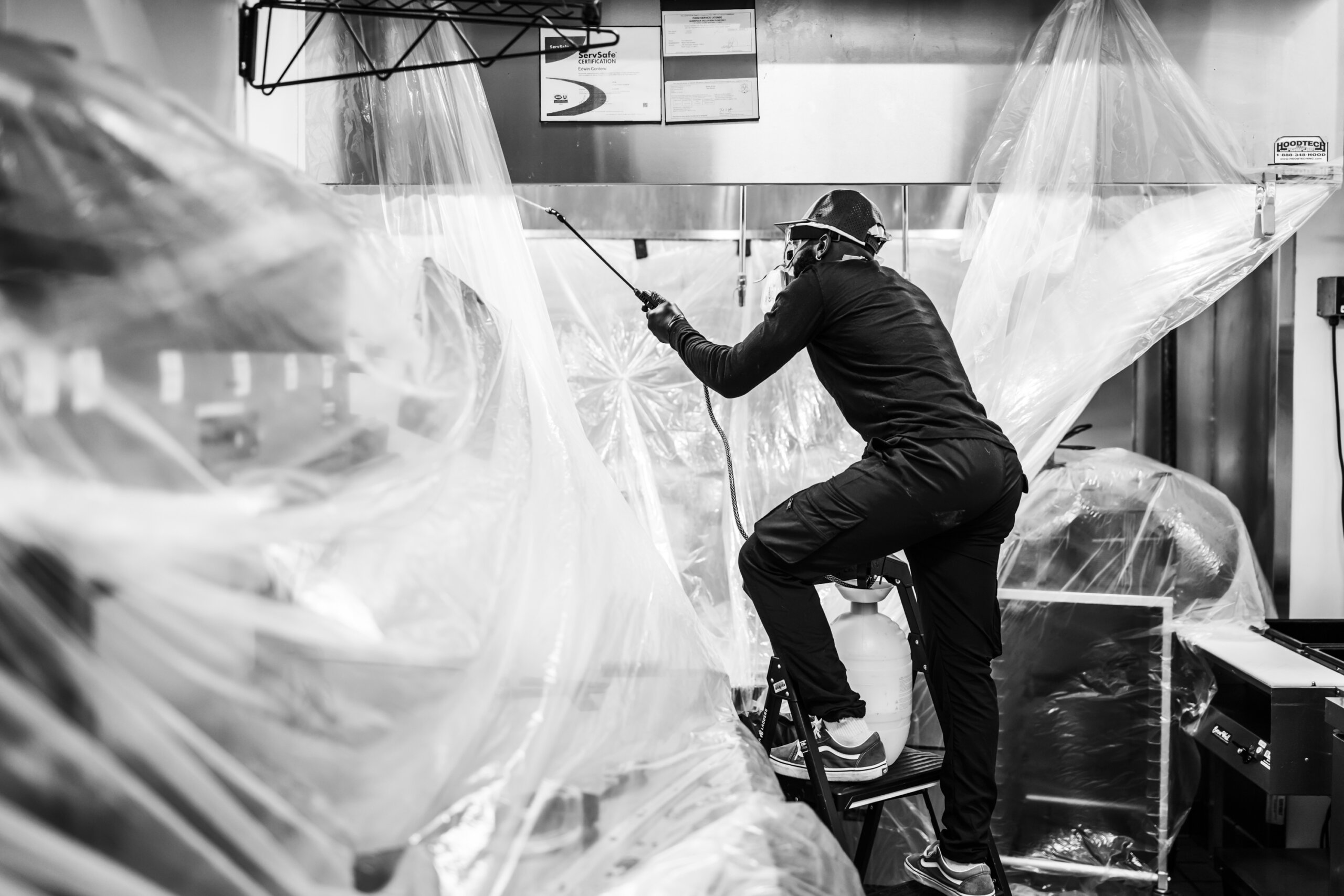 A person stands on a step ladder, spraying liquid in a kitchen area covered with plastic sheets for cleaning or maintenance.