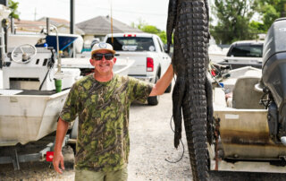 A man in camouflage clothing stands outdoors, smiling and holding up a large, suspended alligator with its snout taped shut; boats and vehicles are visible in the background.