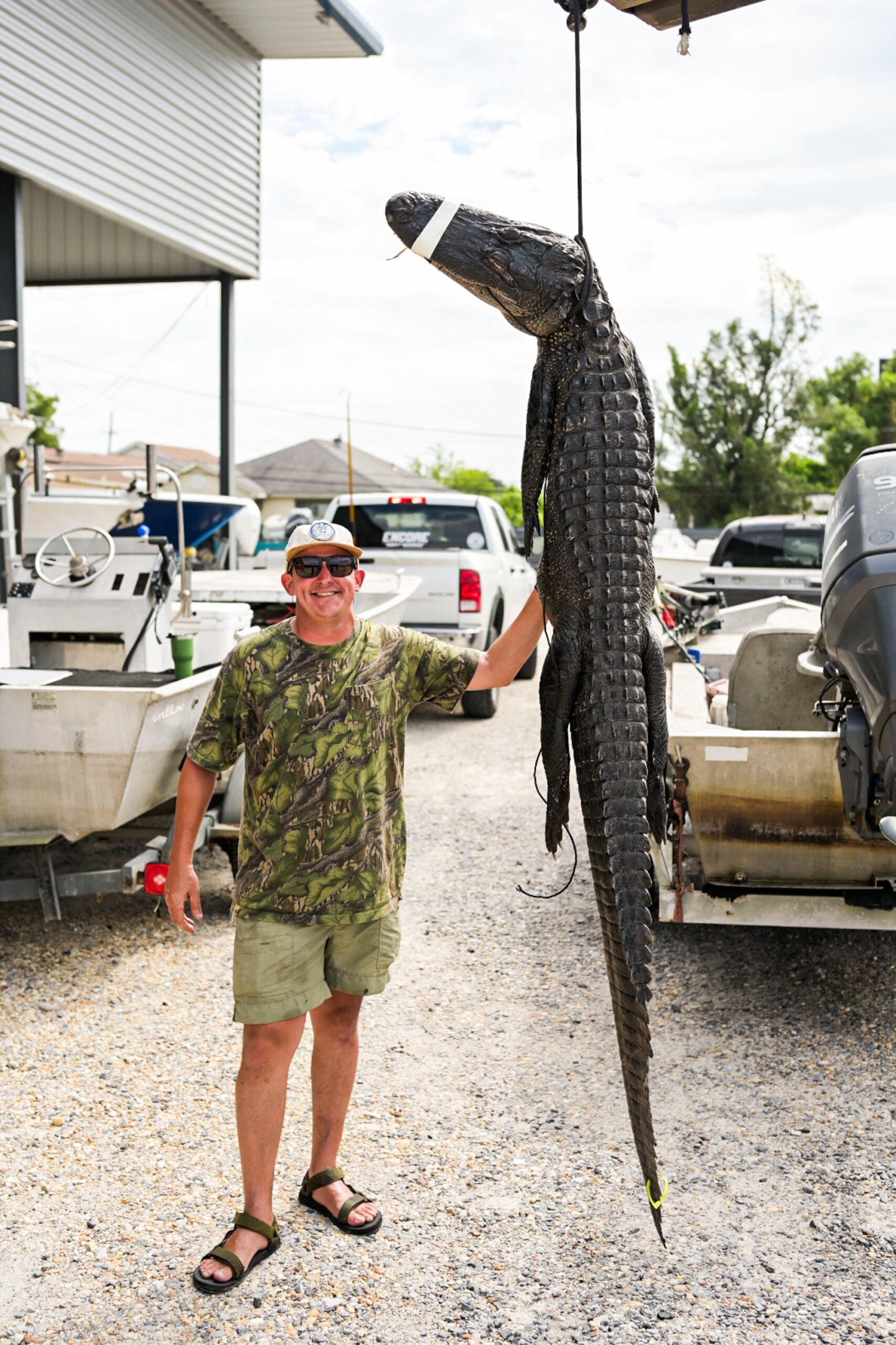 A man in camouflage clothing stands outdoors, smiling and holding up a large, suspended alligator with its snout taped shut; boats and vehicles are visible in the background.