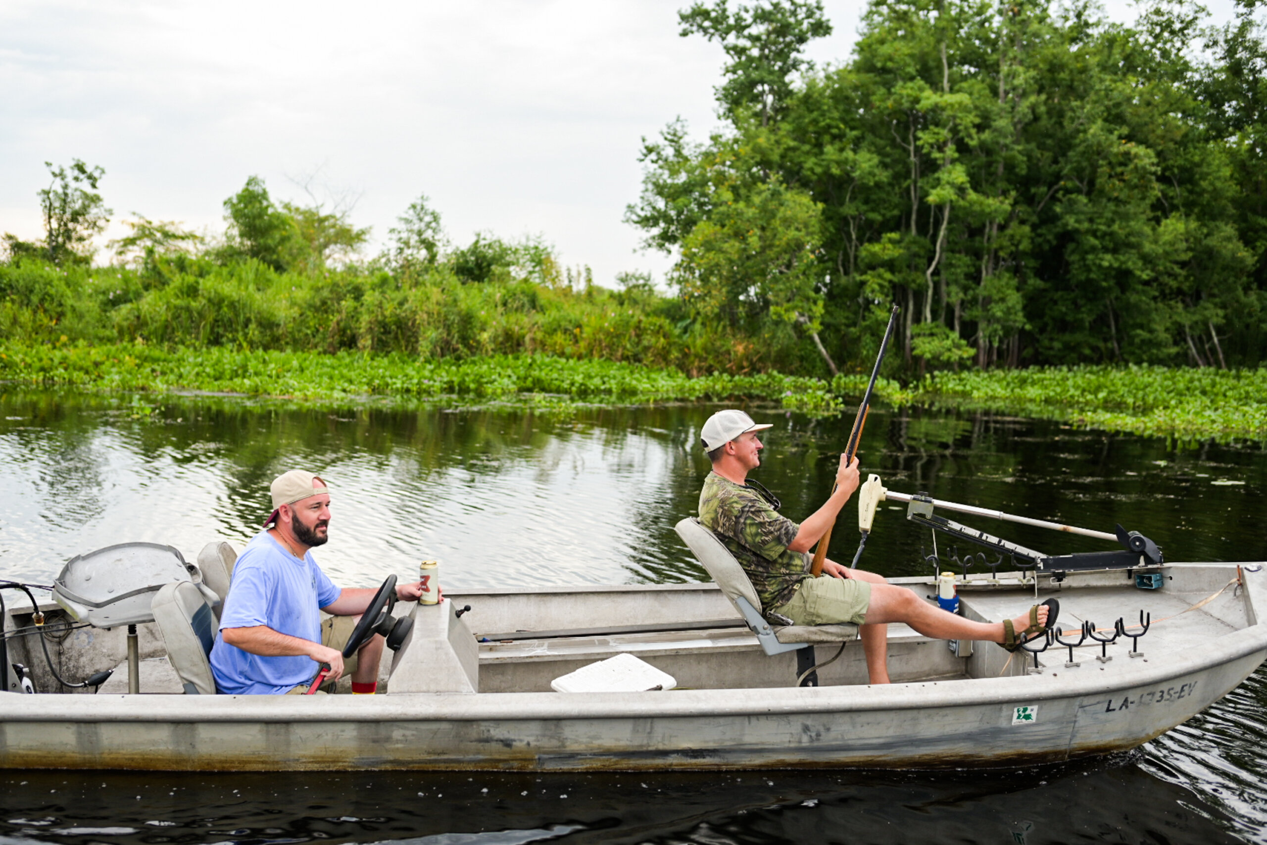 Two men sit in a small motorboat on a calm river; one man steers while the other prepares a hunting rifle. Green trees and plants line the bank in the background.