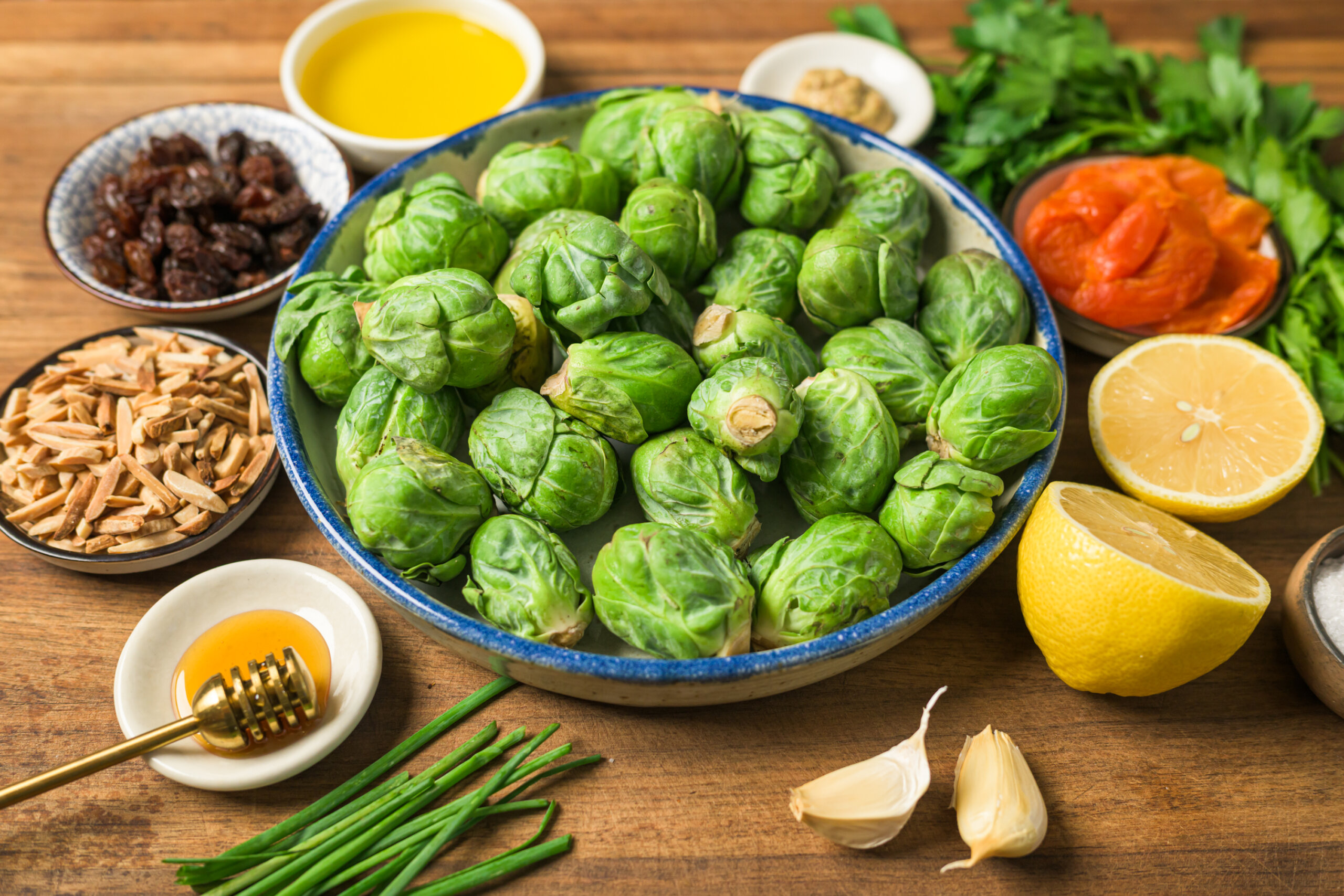 A bowl of fresh Brussels sprouts surrounded by lemon halves, garlic cloves, olive oil, honey, raisins, slivered almonds, herbs, and roasted red peppers on a wooden surface.