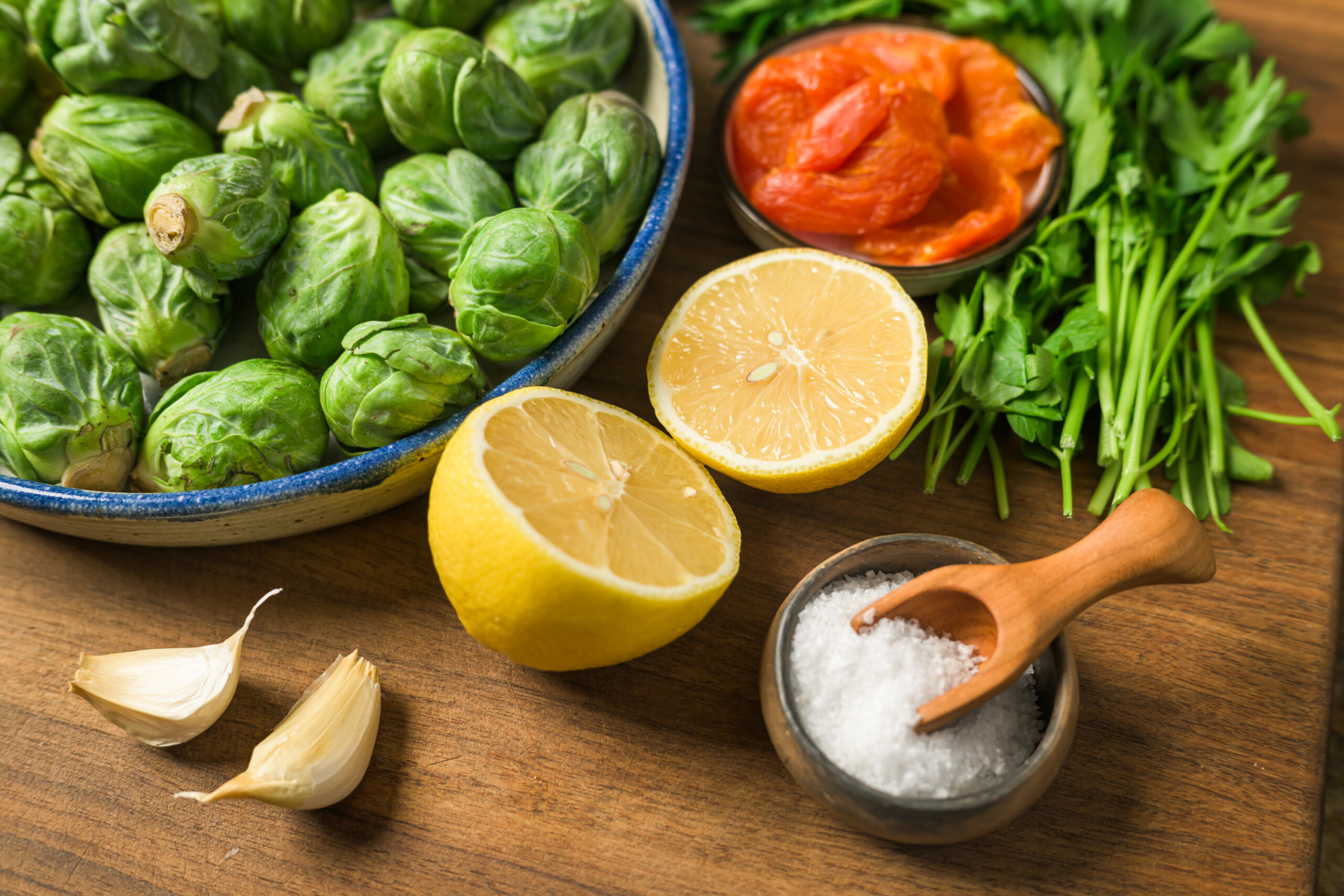 A bowl of Brussels sprouts, halved lemon, salt, garlic cloves, cooked tomatoes, and fresh parsley arranged on a wooden surface.