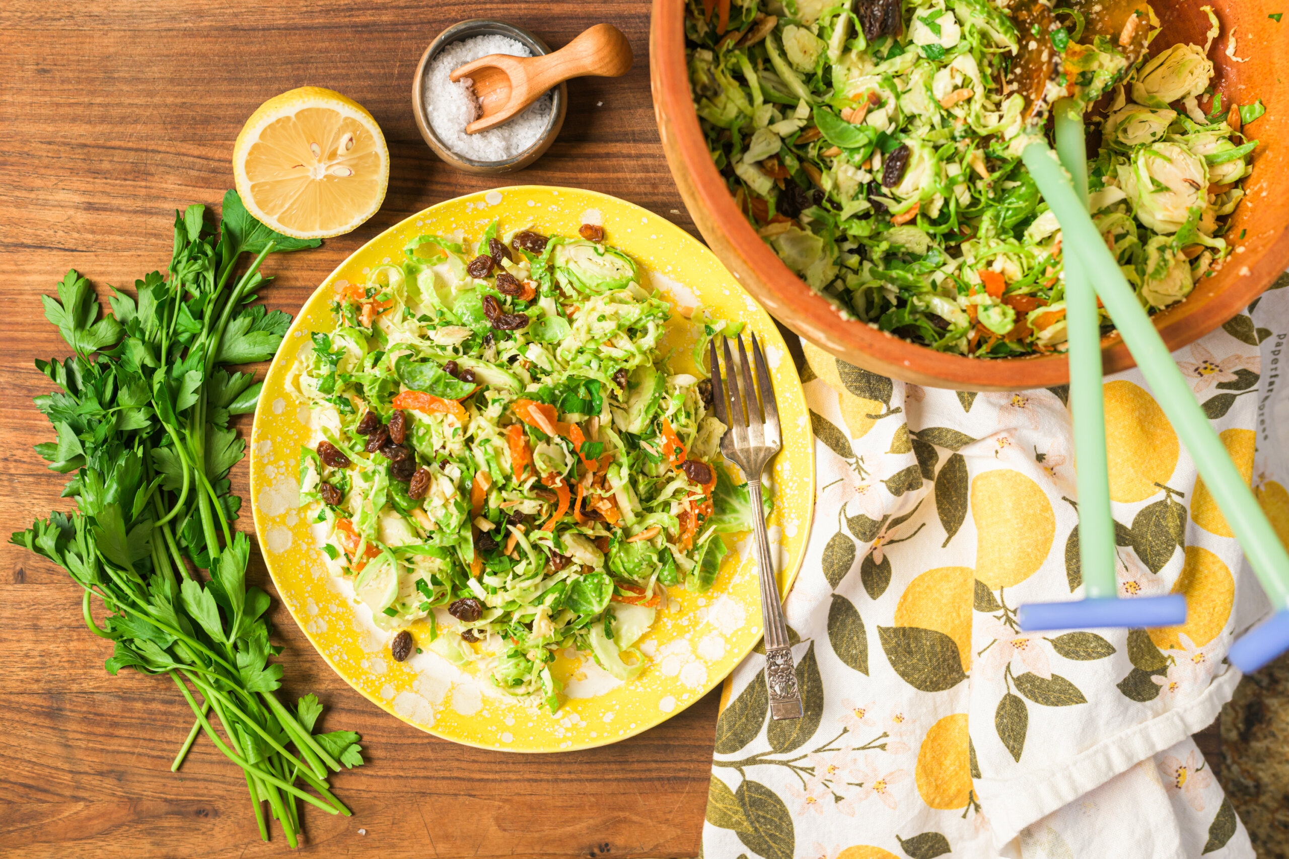 A plate of shredded vegetable salad with parsley, raisins, and carrots on a wooden table, next to a halved lemon, salt bowl, parsley, and a salad bowl with serving tongs.