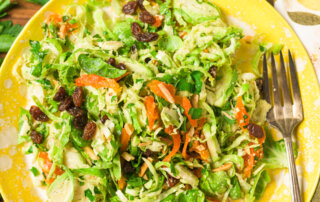 A yellow plate filled with shaved Brussels sprouts salad with parsley, raisins, and dried apricots on a wooden table, next to a halved lemon, salt bowl, parsley, and a salad bowl with serving tongs.