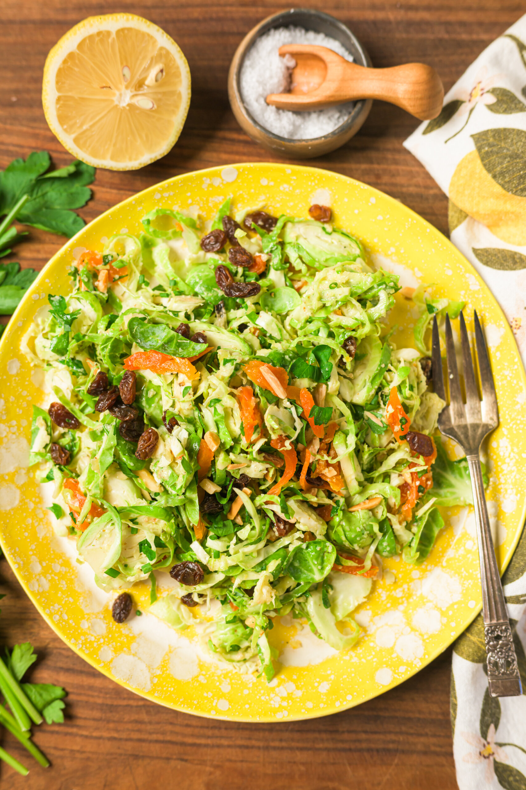 A yellow plate filled with shaved Brussels sprouts salad with parsley, raisins, and dried apricots on a wooden table, next to a halved lemon, salt bowl, parsley, and a salad bowl with serving tongs.