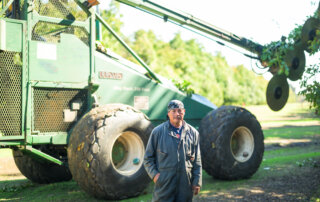 A man in work overalls stands in front of large green agricultural machinery on a sunny day outdoors.