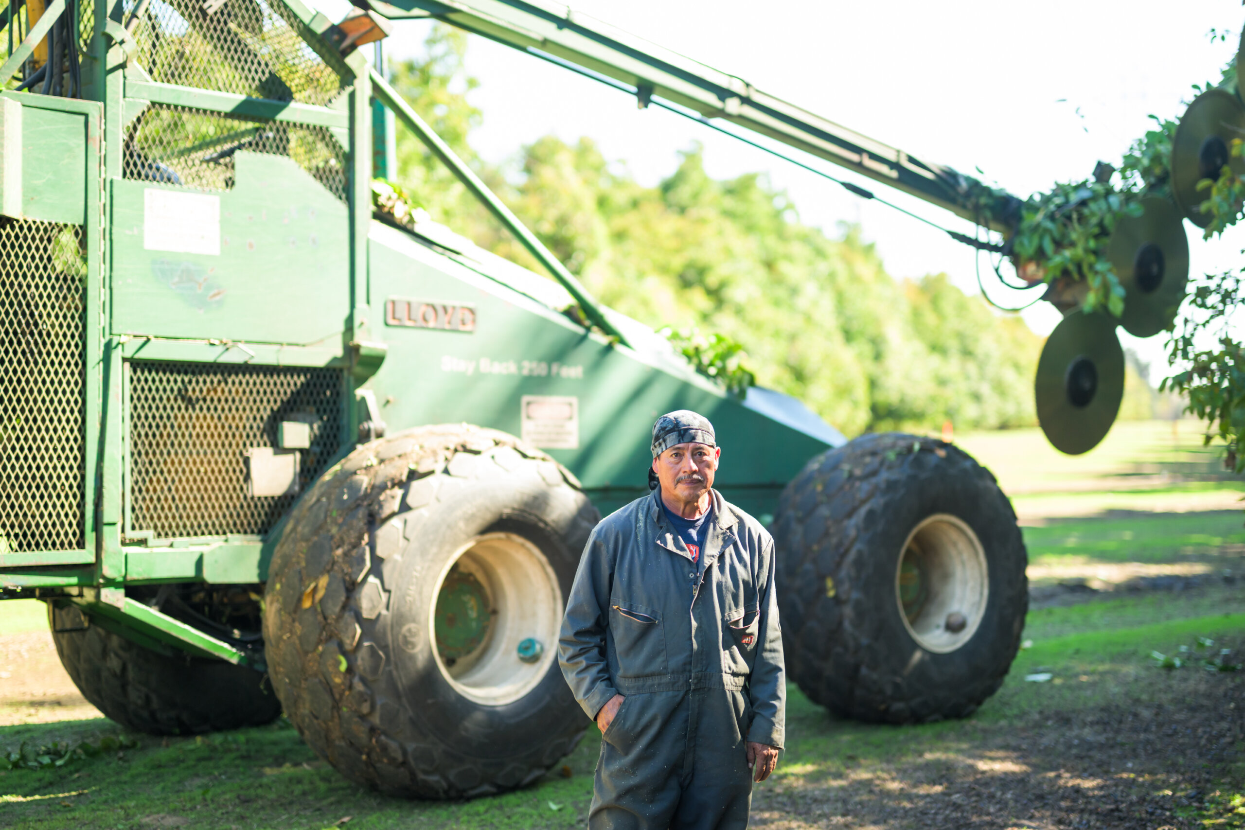 A man in work overalls stands in front of large green agricultural machinery on a sunny day outdoors.