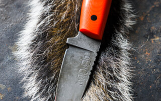 A fixed-blade knife with an orange handle is placed on a strip of animal fur, with the blade marked "DEWCLAW"