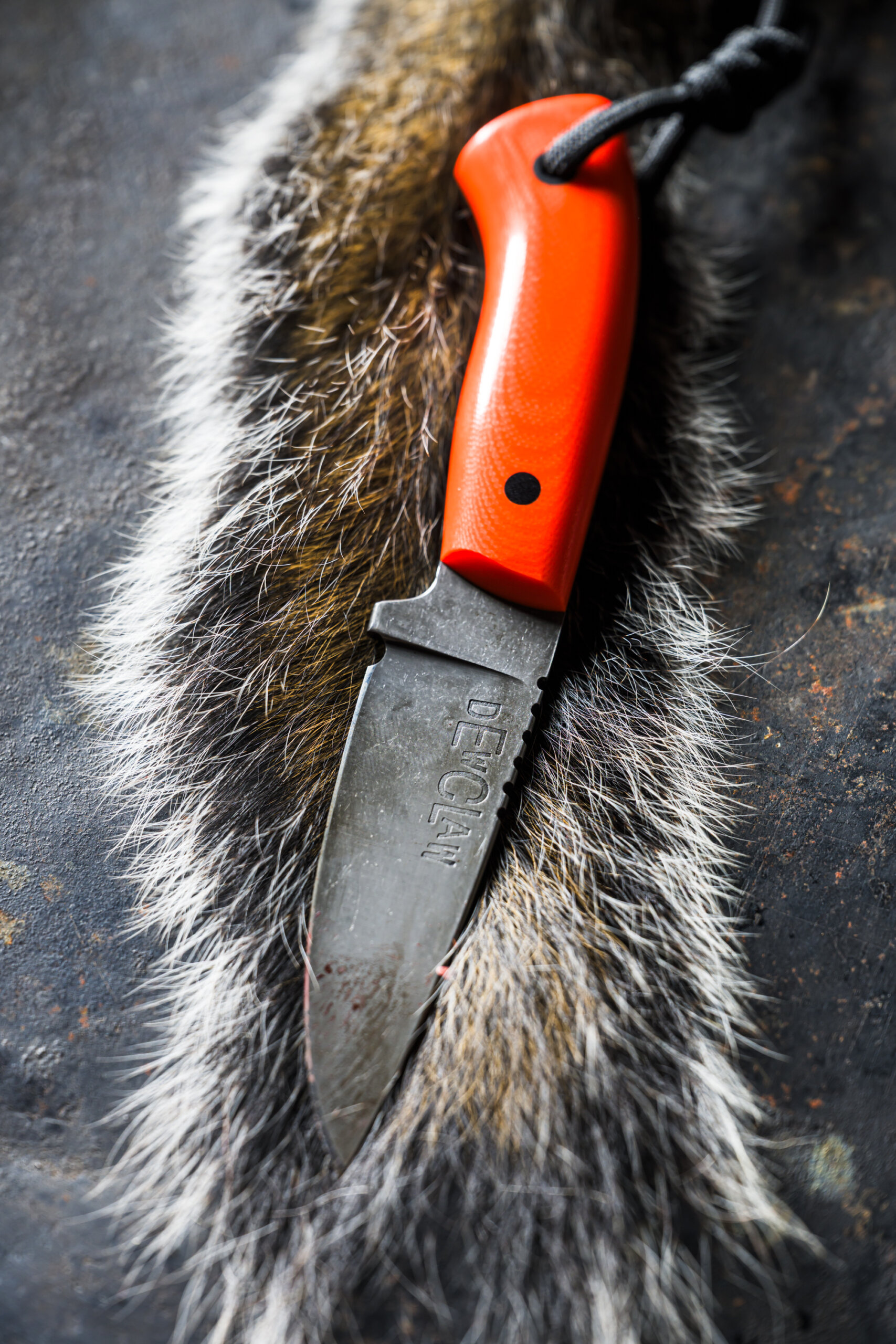 A fixed-blade knife with an orange handle is placed on a strip of animal fur, with the blade marked "DEWCLAW"