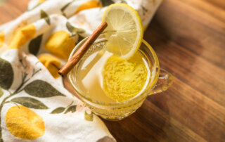 A glass cup of hot lemonade garnished with a lemon slice and a cinnamon stick, placed on a wooden surface beside a lemon-patterned cloth.