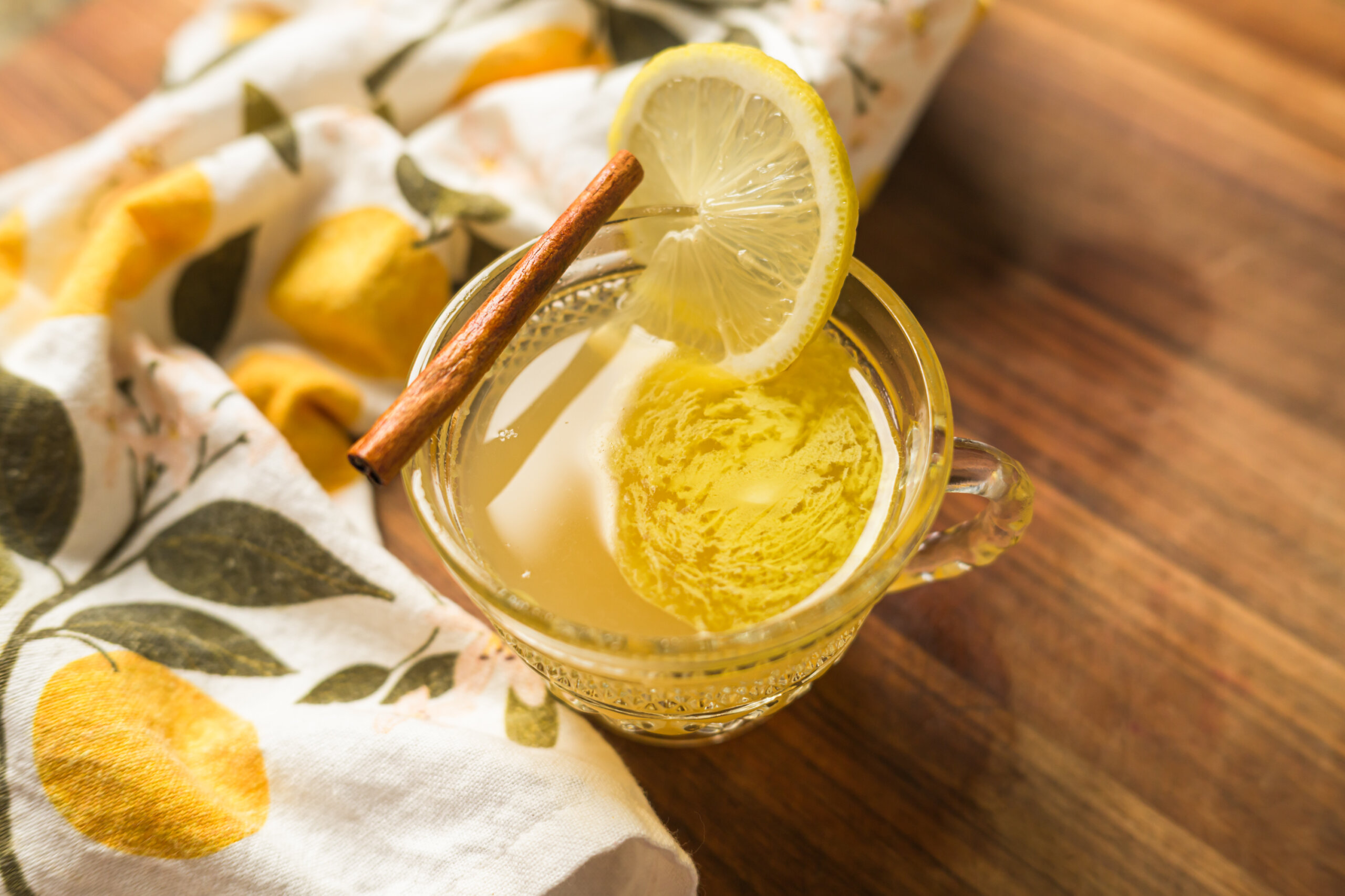 A glass cup of hot lemonade garnished with a lemon slice and a cinnamon stick, placed on a wooden surface beside a lemon-patterned cloth.