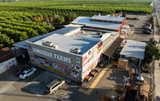 Aerial view of Simonian Farms, with vintage signs on the building, parked cars, farm structures, and orchards in the background under a clear sky.