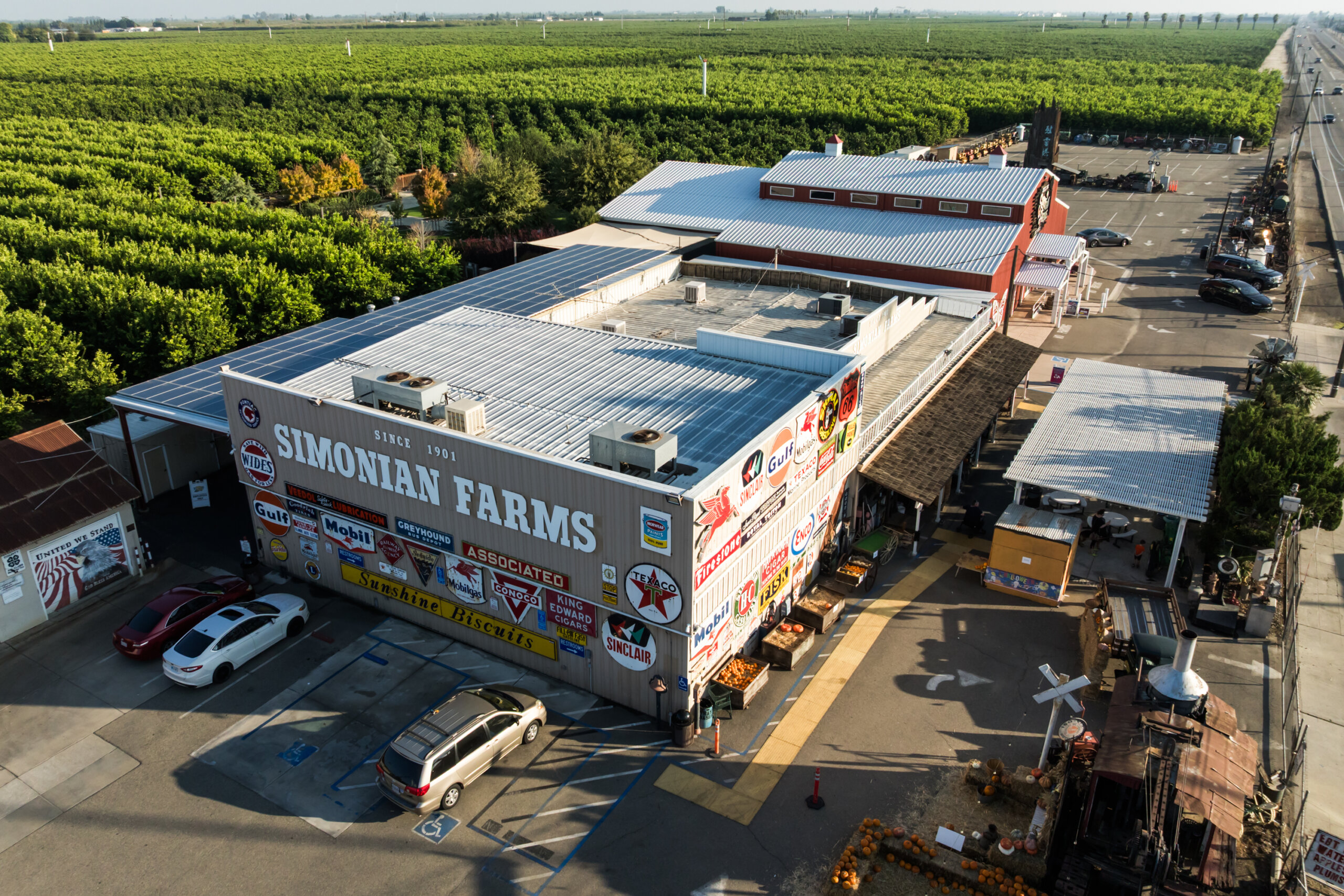 Aerial view of Simonian Farms, with vintage signs on the building, parked cars, farm structures, and orchards in the background under a clear sky.