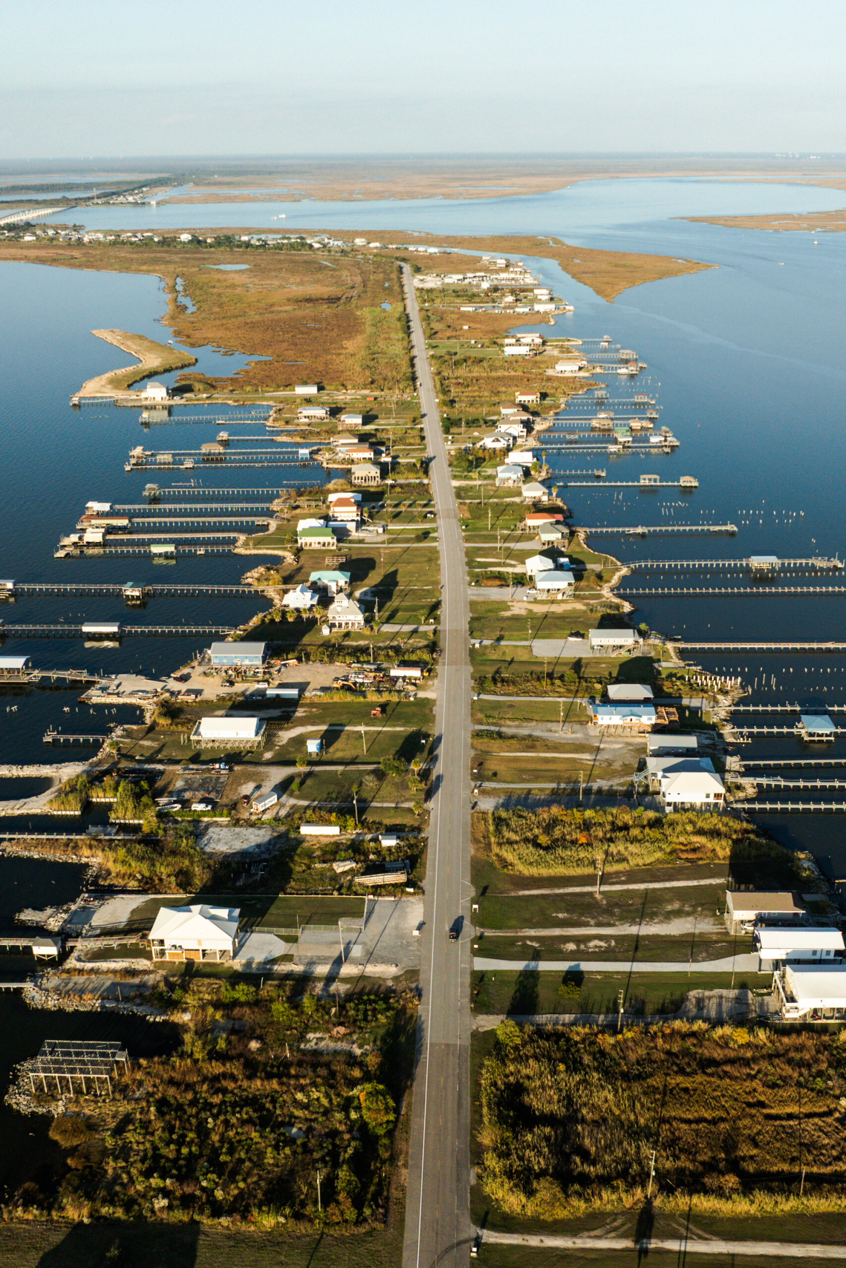 A long, narrow road lined with houses stretches out into a body of water, with docks extending from both sides and marshland visible in the distance.