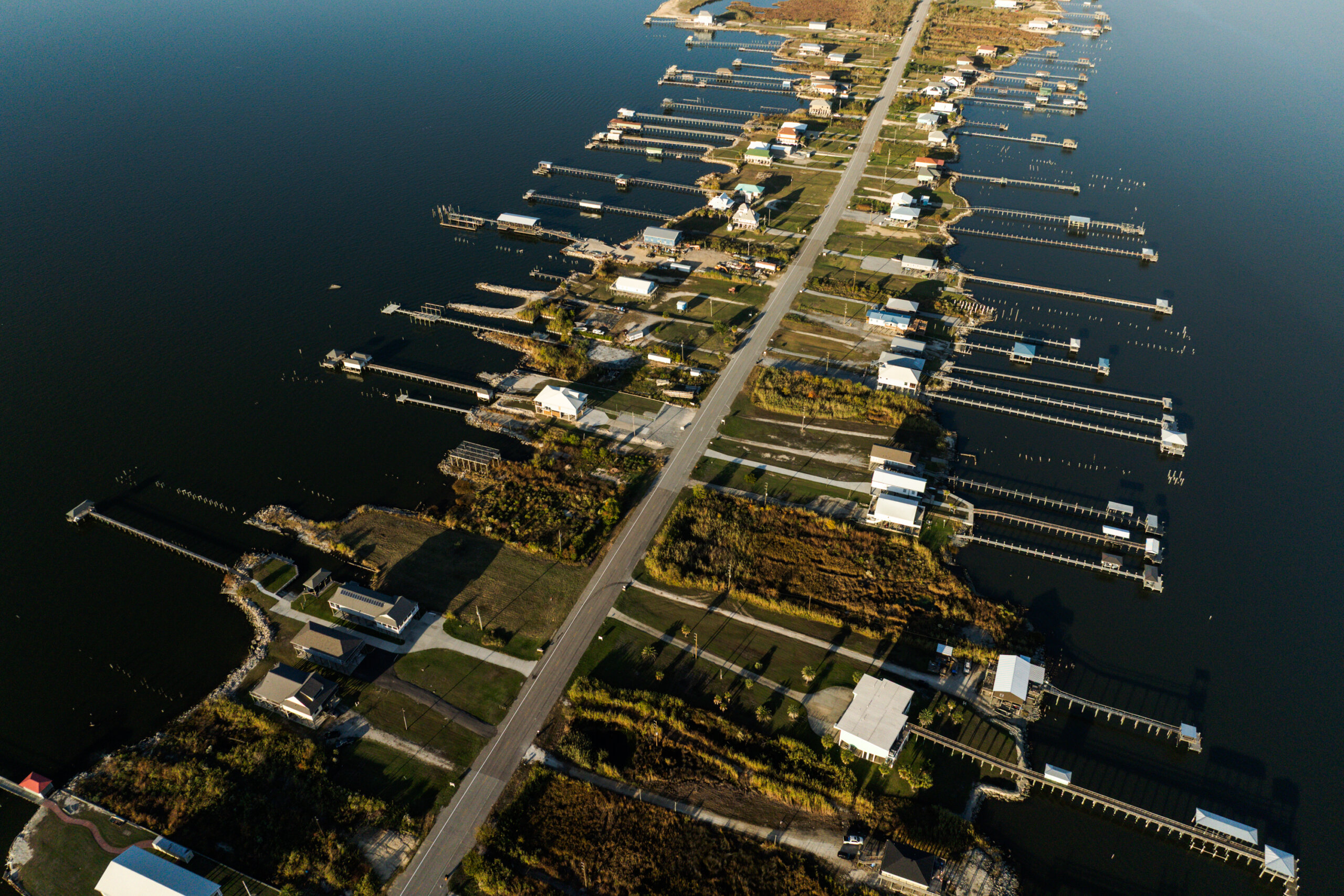 Aerial view of a narrow road with houses and boat docks extending into the water on both sides; land is surrounded by dark water.