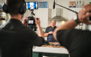 A chef is being filmed in a kitchen by a camera crew. The chef stands behind a counter with cooking ingredients, while microphones and a camera are pointed at him.