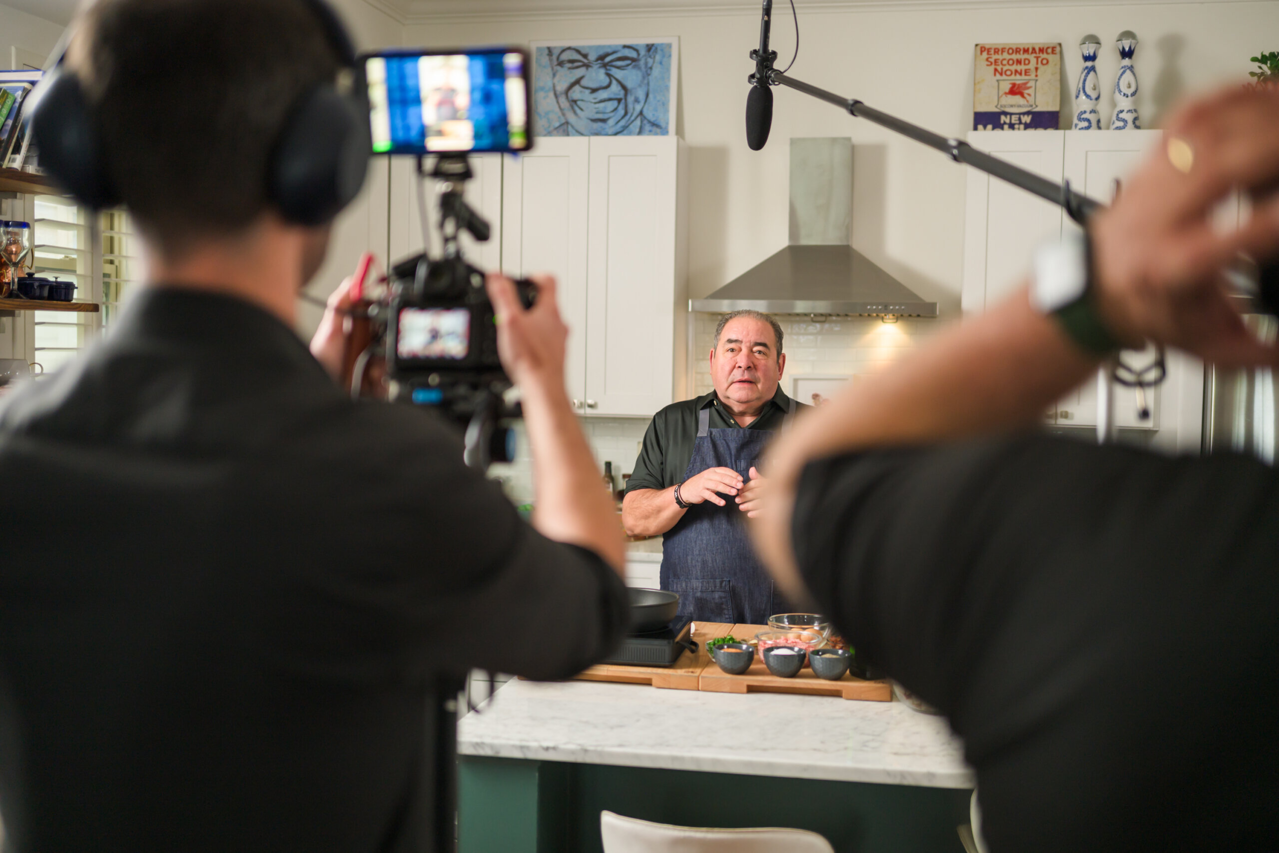 A chef is being filmed in a kitchen by a camera crew. The chef stands behind a counter with cooking ingredients, while microphones and a camera are pointed at him.