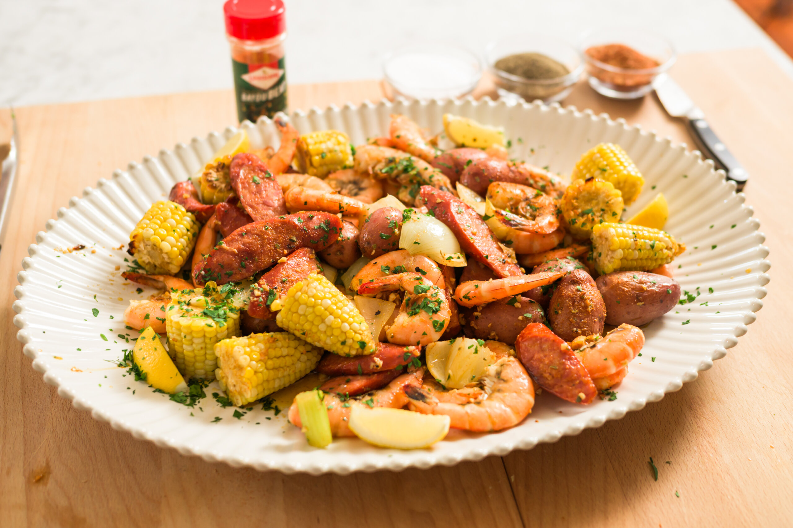 A platter of boiled shrimp, sausage, corn on the cob, potatoes, and lemon wedges garnished with herbs sits on a wooden table with seasonings in the background.