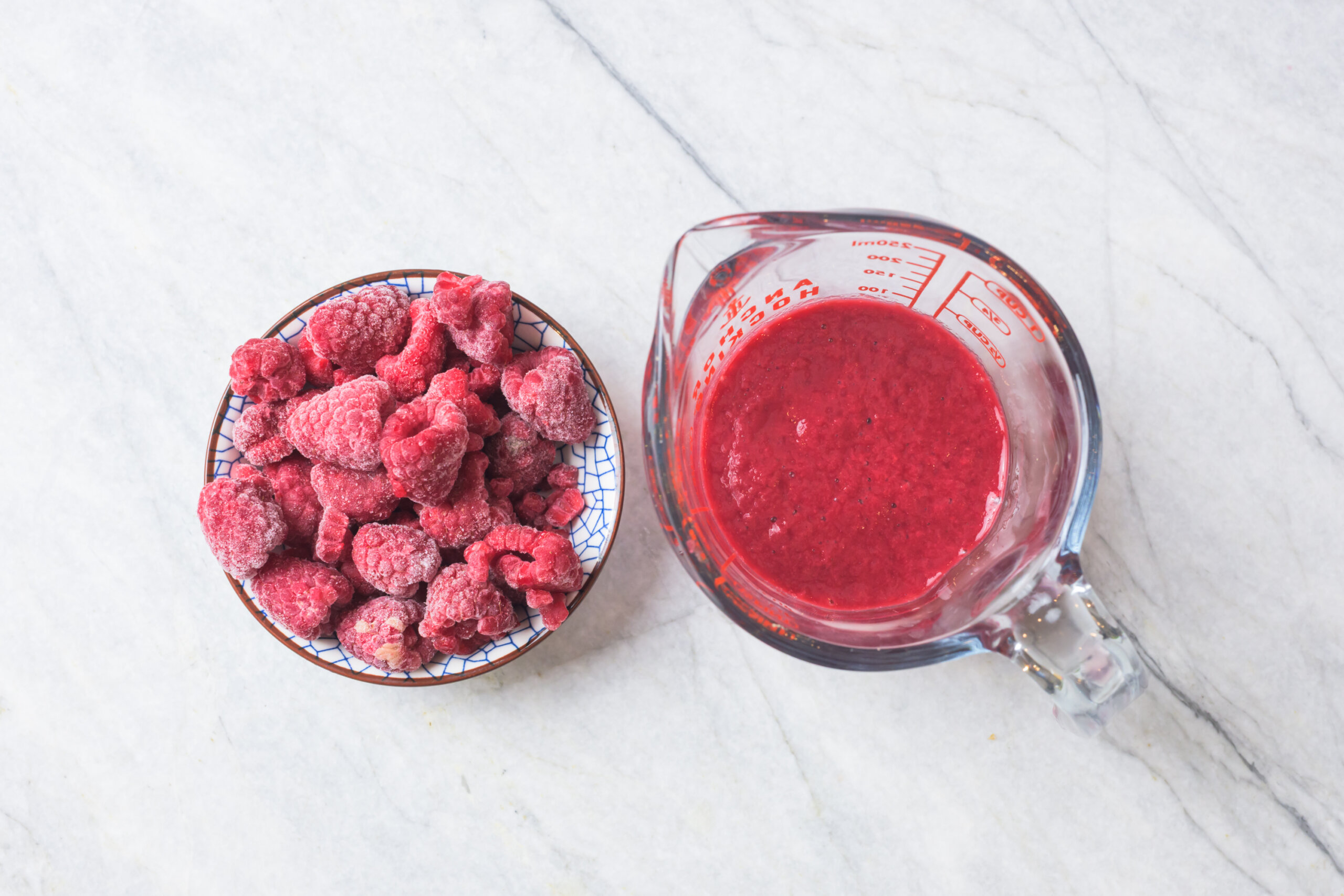 A bowl of frozen raspberries next to a glass measuring cup filled with raspberry puree on a marble countertop.