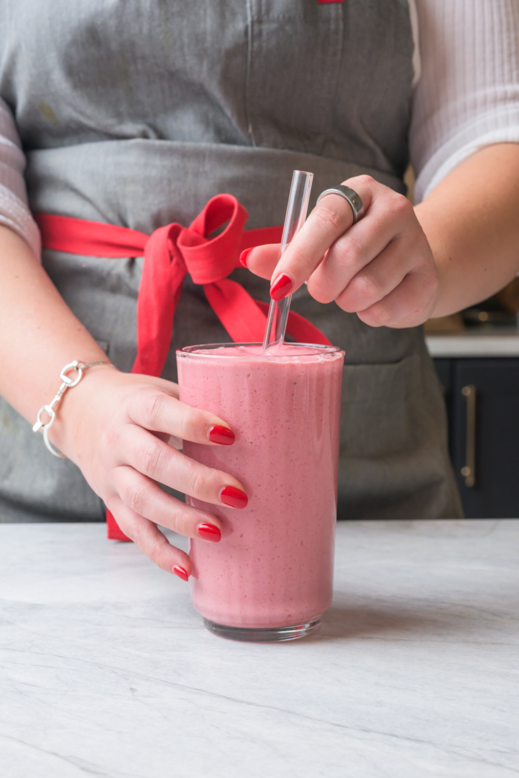 Person in a gray apron with a red tie holding a glass of pink smoothie with a clear straw on a light countertop.